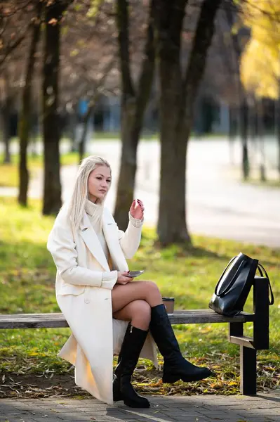 Bir bankta tesadüfen oturan, şık beyaz ceketli bir kadın, sakin bir şehir parkında, ağaçların arasından süzülen güneş ışığına hayran kalarak içkisini yudumluyor..