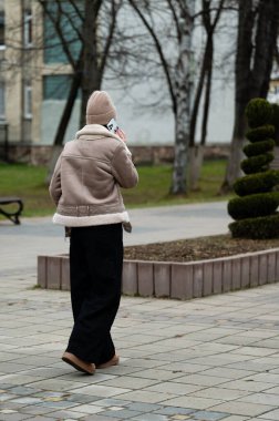 Bir kadın baharın başında sakin bir parkta yürürken, kalın bir ceket ve şapka giyer, görkemli ağaçların ve budanmış çalıların arasında telefon görüşmesi yapar..