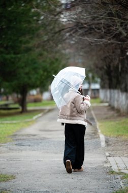 Çürük ağaçlarla çevrili şeffaf bir şemsiyeyle ve bulutlu gökyüzünün altında sakin bir atmosferle huzurlu bir parkta gezinen bir insan..