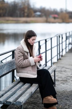 Bu huzurlu an, sakin bir gölün kenarındaki bankta yalnız başına oturan ve doğanın güzelliği içindeki akıllı telefonuna odaklanmış, kalın giyinmiş genç bir kadını yakalıyor..