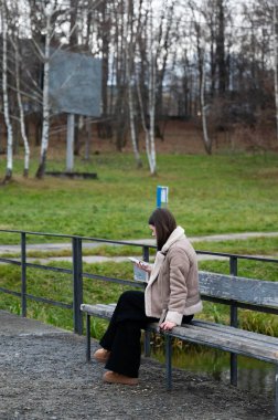 Genç bir kadın bir bankta tek başına oturuyor, bir kitaba dalmış, çıplak ağaçlar ve bir park yolu ile serin bir günde huzurlu bir zemin oluşturuyor, yansımayı ve sakinliği davet ediyor..