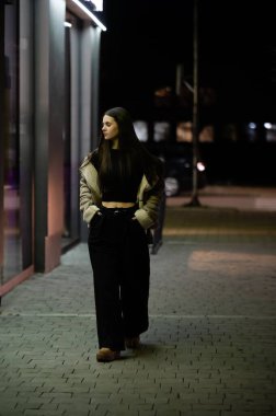 Genç bir kadın şehrin kaldırımında sakin sakin bir şekilde yürüyor, yumuşak şehir ışıkları ve serin gece havasıyla çevrili, sakin ama canlı bir ortam yaratıyor..