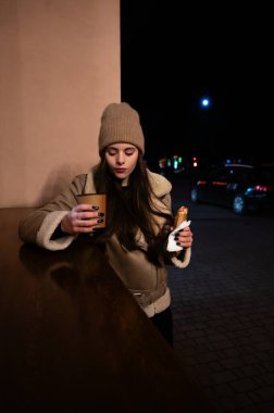 Genç bir kadın gecenin bir yarısı ahşap bir tezgahın yanında durup elinde lezzetli bir aperatif tutarken sıcak bir içeceğin tadını çıkarıyor..