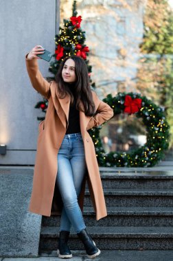 Genç bir kadın güvenli bir şekilde merdivenlerde duruyor, şık bir şekilde giyinmiş, şık bir şehir ortamında tatil süslemeleriyle çevrili..