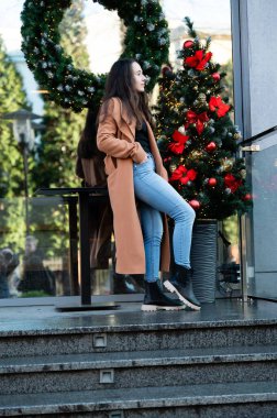 Şık bir ceket giymiş, kendine güvenen bir kadın kış gününün tadını çıkarır. Parlak süslemelerle süslenmiş bir Noel ağacının yanındaki korkuluklara yaslanır..