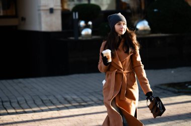 Parlak güneş ışığı, şık bir kadının üzerinde parıldıyor. Şehir caddesinde rahatça geziniyor, sıcak bir içeceğin tadını çıkarıyor ve şık kış kıyafetini sergiliyor..