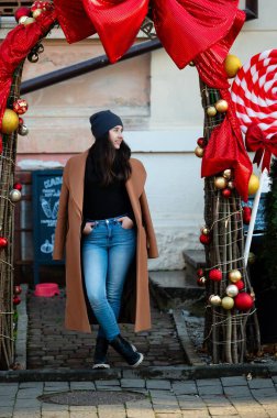 Zarif genç bir kadın zarif bir şekilde güzel dekore edilmiş bir kemer yolunun altında duruyor. Etrafı renkli tatil süsleriyle çevrili. Kış gününde neşe saçıyor..