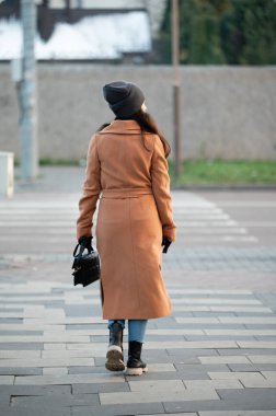 Bir kadın güvenli bir şekilde kaldırımda yürüyor, bronz bir trençkot giyiyor ve rahat bir bere takıyor, şık yeteneğini sergiliyor ve güzel bir sonbahar gününün tadını çıkarıyor..