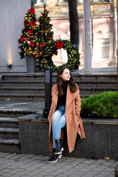 Şık bir palto giymiş genç bir kadın, bir bankta rahat bir şekilde oturur, güzel bir tatil süsü alır ve kış soğuğunun ortasında onu çevreleyen parlak ışıkları yakar..