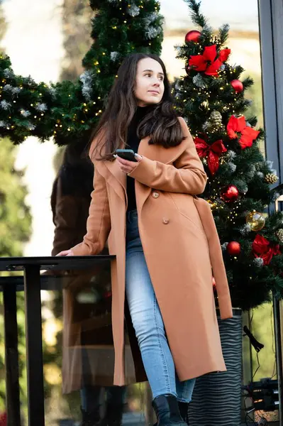 Uzun saçlı genç bir kadın, parlak süslemelerle süslenmiş bir Noel ağacının yanında gelişigüzel duruyor. Şehir ortamında kış havasının tadını çıkarıyor..