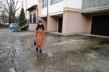 Genç bir kadın şık bir kahverengi ceket içinde sakin bir şekilde yürümekten hoşlanır. Serin bir günde sakin bir kentsel alanın kaldırımında su birikintilerinde gezinirken kış şapkası.