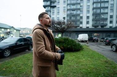 Sıcak bir şekilde giyinmiş bir adam, akşamları modern bir mahallede duruyor. Etrafı park edilmiş arabalar ve modern binalarla çevrili iken manzarayı seyrediyor..