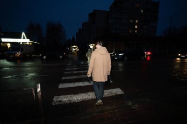 Sakin bir kentsel alanda yağmurlu bir akşamda yavaşça ilerleyen araçlarla çevrili, sönük sokak lambalarının altında, yaya geçidinde güvenle yürüyen bir insan..