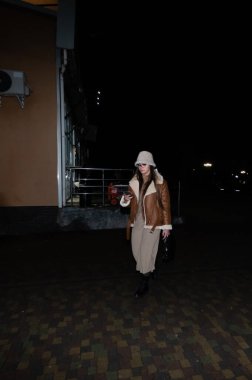 Sıcak kahverengi bir ceket ve açık renk bir elbise giymiş şık bir birey yakındaki ışıkların altında hareketli bir caddede rahatça dolaşır zarafet ve zarafet saçar.