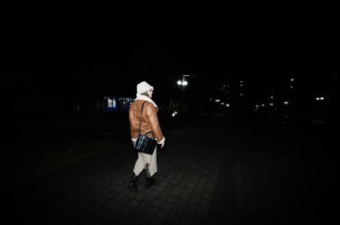 Sokak lambalarının parıltısı altında, kahverengi deri ceketli ve beyaz kapüşonlu bir insan kaldırım taşı yolda yürüyor, bir gece şehir atmosferinin sakinliğini kucaklıyor..