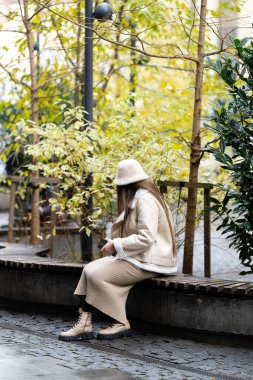 Şık bej giysiler giymiş bir kadın sakin bir şehir parkında yemyeşil bir bankta dinleniyor ve öğleden sonra sakin bir atmosferi kucaklıyor..