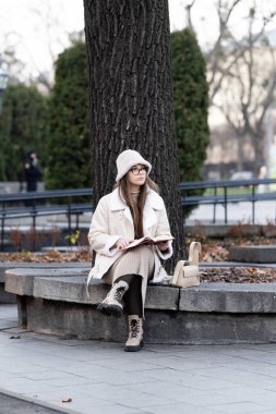 Sıcak tabakalara sarılmış genç bir kadın, bir sonbahar öğleden sonrasının huzurlu atmosferinde, büyük bir ağacın altındaki taş bir bankta oturuyor..