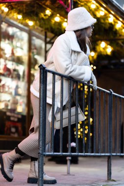 Sıcak bir kışlık palto ve şapka giymiş bir kadın, canlı bir şehir noktasında ışıl ışıl tatil ışıklarıyla çevrili bir korkuluğa yaslanıyor ve sakin bir mevsim havası yaratıyor..