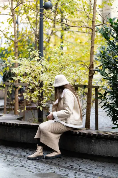 Şık bej giysiler giymiş bir kadın sakin bir şehir parkında yemyeşil bir bankta dinleniyor ve öğleden sonra sakin bir atmosferi kucaklıyor..