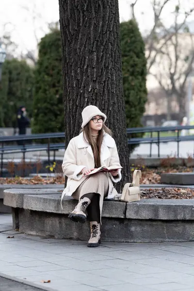 Sıcak tabakalara sarılmış genç bir kadın, bir sonbahar öğleden sonrasının huzurlu atmosferinde, büyük bir ağacın altındaki taş bir bankta oturuyor..