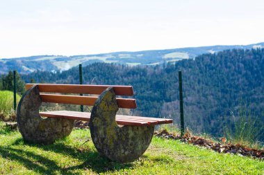 Benzersiz tasarlanmış bir bank çimenli bir tepecikte oturur ve ziyaretçileri parlak mavi gökyüzünün altında yuvarlanan yeşil tepelerin geniş manzarasının tadını çıkarmaya davet eder..