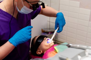 Temiz ve renkli bir diş kliniğinde, önlüklü bir diş hekimi genç bir hastayla dikkatlice ilgilenir. Çocuk koruyucu gözlük takıyor ve ameliyat sırasında sakin görünüyor..