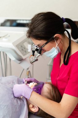 İlgili bir dişçi, parlak ve misafirperver bir diş kliniğinde genç bir çocuğun dişlerini modern aletlerle muayene eder. Çocuk rahat görünüyor, özen ve destek görüyor..