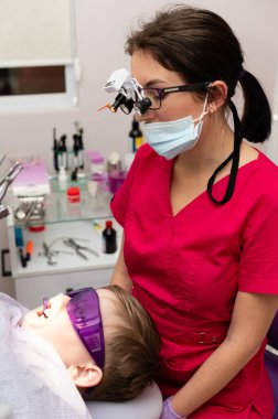 Canlı üniformalı bir diş hekimi, özel bir gözlük giyerken genç bir hastayı dikkatlice muayene eder ve pediatri kliniğinde rahat bir deneyim sağlar..