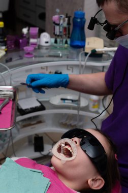 Bir dişçi dişlerini incelerken, küçük bir çocuk diş sandalyesinde koruyucu gözlük takıyor. Klinik modern aletler ve canlı renklerle donatılmıştır..