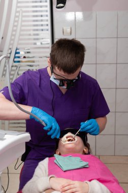 Diş hekimi muayenesi yaparken, hasta dişçi koltuğunda dinleniyor. Rutin kontroller sırasında, parlak ve modern bir klinik ortamında oral bakım araçları kullanıyor..