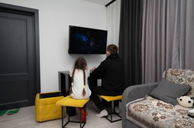 İki kişi, şık bir oturma odasında, rahat bir dekorla çevrili, televizyonda ilgi çekici bir film izlerken, parlak sarı taburelerde otururlar..