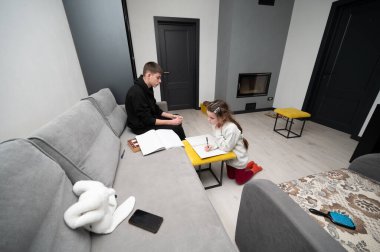 İki kardeş modern bir oturma odasında ders çalışmaya odaklanıyor. Bir çocuk yerde oturmuş, diğeriyse derin düşüncelere dalmış bir şekilde koltukta oturuyor..