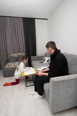 Genç bir kız çizimlerine derinden odaklanarak yerde otururken, kanepede oturan bir genç notları inceleyerek öğrenme ve iletişim kurma konusunda sıcak bir ortam yaratıyor..