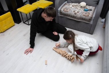 İki genç, sıcak bir oturma odasındaki hafif bir zemine ahşap blokları yığmaya odaklanmış, rahat bir atmosferin ortasındaki oyuncu yaratıcılıklarını ve takım çalışmalarını ön plana çıkarıyorlar..