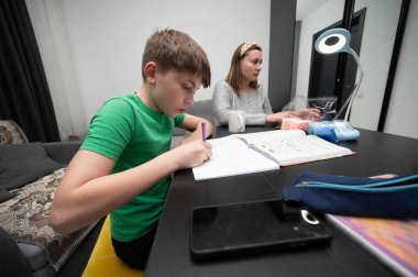 Genç bir çocuk, evde destekleyici bir öğrenme ortamı yaratarak bir tartışmayla meşgul olan annesinin yanında otururken ev ödevine dikkatle odaklanır..