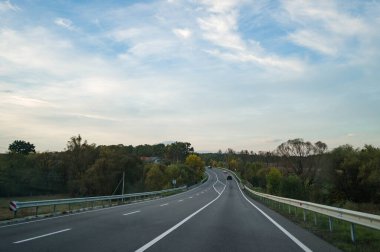 İleride, canlı sonbahar renklerini gösteren ağaçlarla çerçevelenmiş kıvrımlı bir yol uzanıyor. Huzurlu gökyüzü huzurlu atmosfere katılıyor ve kırsal bölgelerin keşfine davet ediliyor..