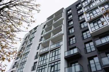 Modern çok katlı bir apartmanın çarpıcı manzarası, sonbahar yaprakları ve bulutlu gökyüzü ile çevrili, parlak hatlar ve geniş balkonları ön plana çıkarıyor..