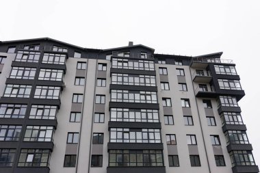 Geniş pencereleri ve parlak bir cephesi olan çağdaş bir yerleşim yeri gri gökyüzüne karşı duruyor, modern kentsel yaşam ve mimari yenilikler sergiliyor..