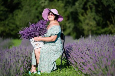 Çiçek açan lavanta çiçeklerinin arasında oturan bir kadın, elinde mis kokulu mor çiçeklerden oluşan bir sepet tutuyor. Bir yaz elbisesi ve geniş kenarlı bir şapka takıyor, tarlanın huzurlu güzelliğinin tadını çıkarıyor..