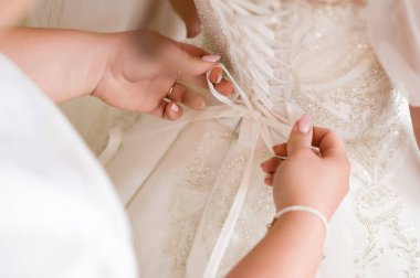 Zarif beyaz bir gelinliğin bağcıklarını sıkıca bağlayan iki elin yakın çekimi. Elbise çok detaylı ve iyi bir işçiliği var..