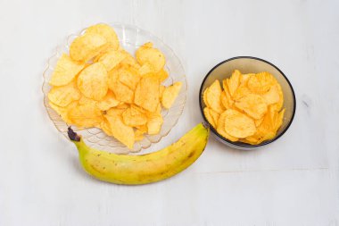 Çıtır altın muz cipsleri, beyaz ahşap bir masanın üzerinde taze, olgun bir muzla birlikte, küçük, siyah bir kasede servis edilir..