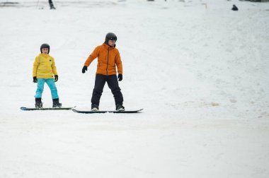 Canlı mavi kıyafetli bir çocuk ve turuncu giyinmiş bir yetişkin, karlı bir yamaçta kar kayağı yapmaktan zevk alıyor. Açık havada kış sporlarının keyfini çıkarıyorlar..