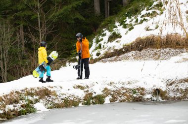 İki arkadaş, kısmen donmuş bir göletin kenarında durmuş kar aktiviteleriyle dolu maceralı bir gün için heyecanlı bir şekilde snowboard hazırlıyorlar..