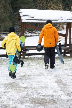 Kış kıyafetleri içinde iki kişi kar kayaklarıyla yamaçlara doğru yürüyorlar. Snowboard ve macera dolu heyecanlı bir güne hazırlar..