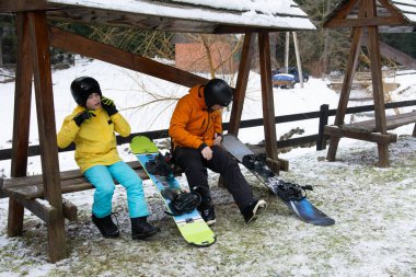Parlak kış giysileri içinde iki genç arkadaş kar kayaklarıyla bir bankta oturup kar kaplı yamaçlarda öğleden sonra eğlencesine hazırlanıyorlar..