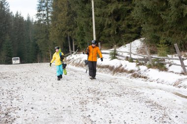 İki arkadaş, parlak kış kıyafetleri içinde, karlı bir yolda yürüyor, açık havada eğlenceli bir güne hazırlanıyorlar, etrafları uzun yeşil ağaçlar ve güzel dağ manzaralarıyla çevrili..