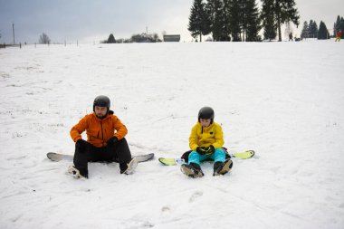 İki kişi, karlı bir arazide kızaklara binip açık hava eğlenceleriyle dolu bir kış gününde dinleniyorlar. Ağaçlar arka planda kalır, dingin atmosfere eklenir.