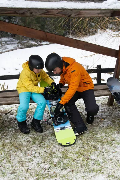 Küçük bir çocuk, bir yetişkinin snowboard malzemelerini hazırlamasına yardım etmesini hevesle izler..