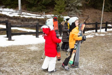 Canlı kayak kıyafetleri içindeki çocuklar kayaklarını yamaçlara doğru taşırlar. Arka planda kar parıldıyor, eğlence ve macera dolu bir günü ima ediyor..