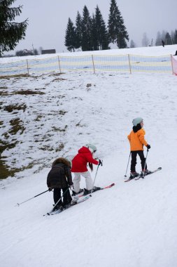Renkli ceketli üç çocuk, sakin kış manzarasının ortasında kayak becerilerini çalışırken karlı bir tepeye doğru yol alıyorlar..